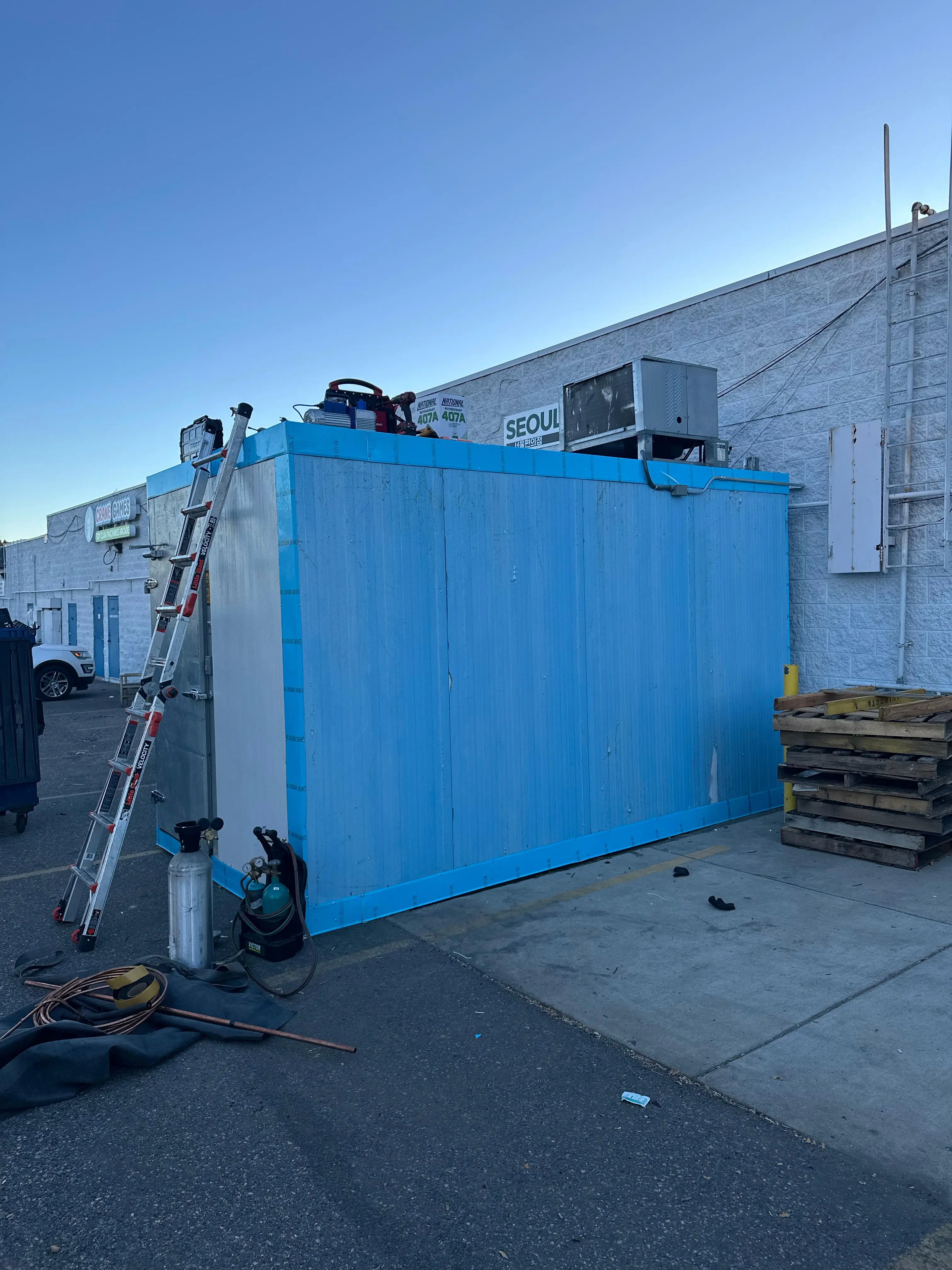 A blue insulated walk-in cooler built by Friantartic behind a shopping center, with a ladder, 407A refrigerant and a coil of copper line