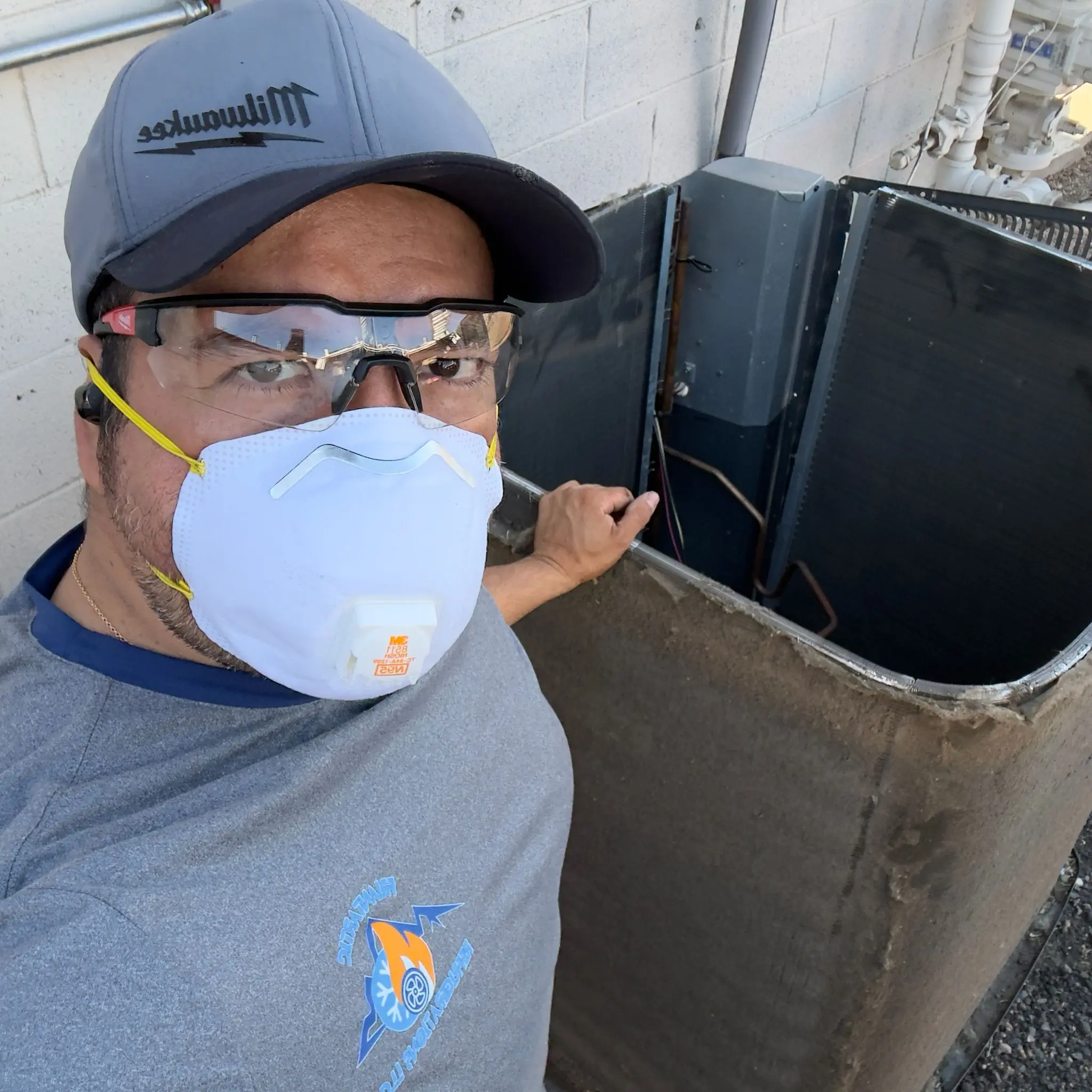 A Friantartic technician in a cap, safety goggles and N95 mask beside a condenser he is cleaning, the company logo on his shirt